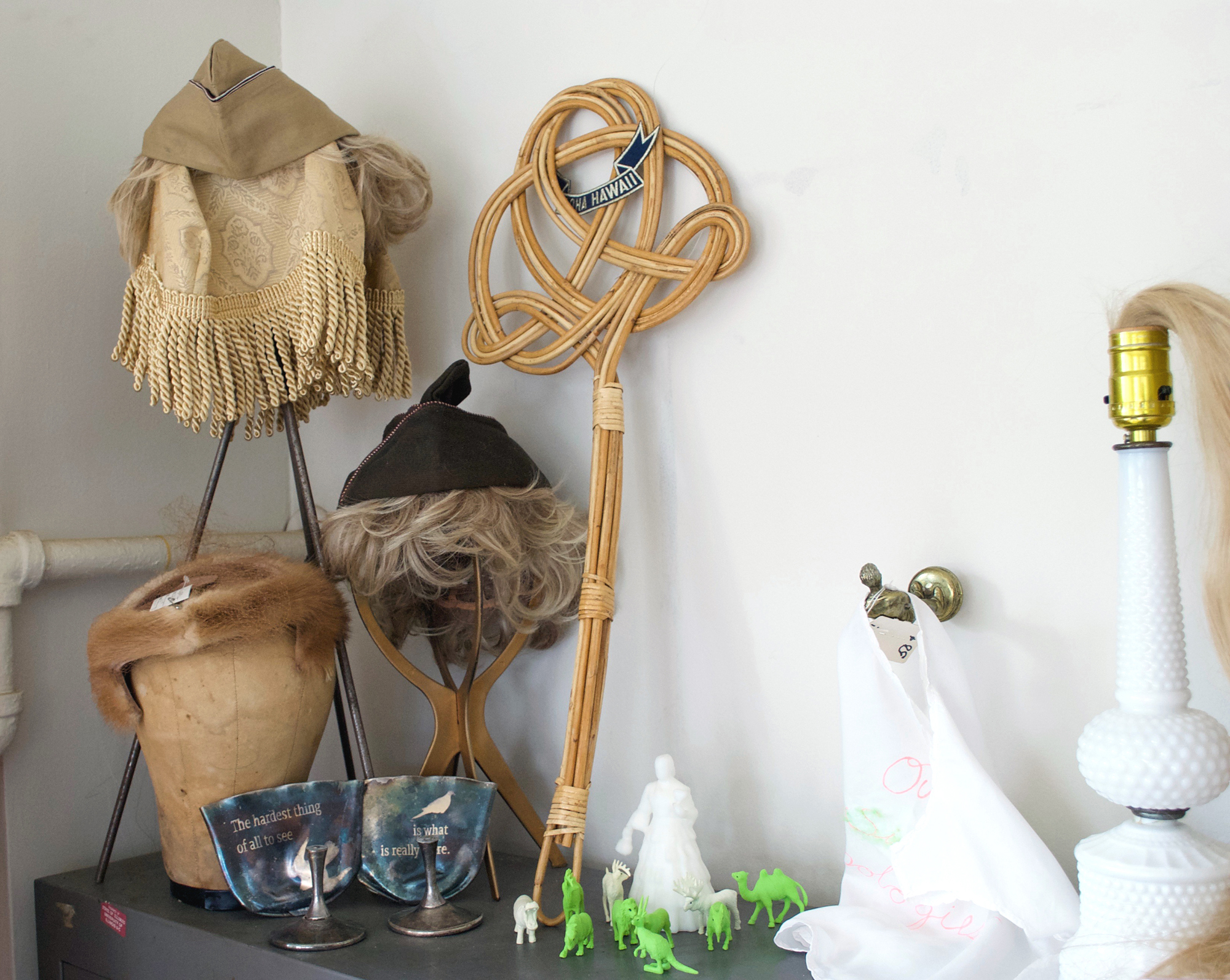 Image: A collection of objects sit on a shelf in Montgomery’s studio. From left to right: a wooden wig head holds a brown fur cap; a metal stand holds a piece of fringed, beige fabric atop which sits a wedged cap with blonde hair; another metal stand holds a wedged cap with blonde hair; a wooden rug beater; small human and animal figurines; a lamp with blonde hair in the place of a lightbulb. Photo by Jessica Hammie.