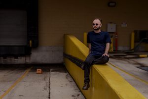 Featured Image: Elliot J. Reichert sits, legs crossed, on a yellow concrete platform in what appears to be the exterior of a loading dock. He's looking out to the left of the frame. Photo by Ryan Edmund.