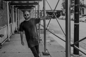 Image: Elliot stands under scaffolding along a brick building, with the street to the right. One hand holds onto a post on the scaffolding, and he's leaning out to the left towards the frame. Photo is in black and white. Photo by Ryan Edmund.