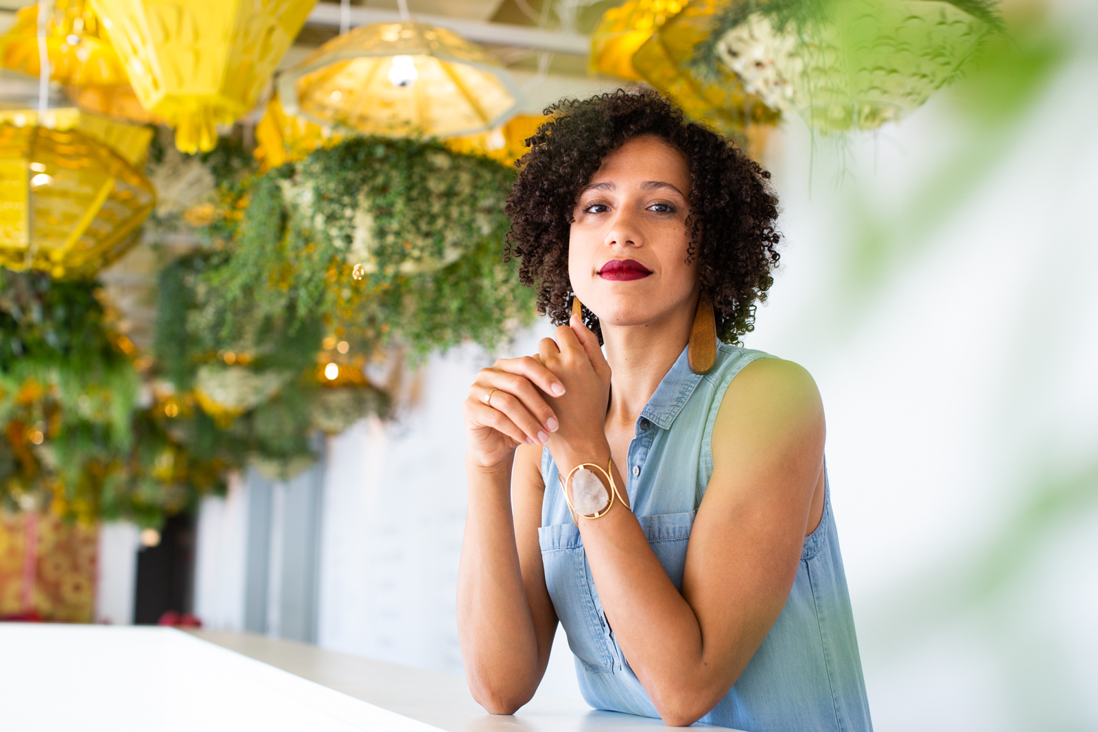 A portrait of Tara Aisha Willis, the Associate Curator of Performance at the Museum of Contemporary Art Chicago. Tara is standing with elbows on a ledge, hands up toward her face and fingers intertwined. She stands in a space where there are plants and golden yellow lights hanging from the ceiling. Photo by Kristie Kahn.