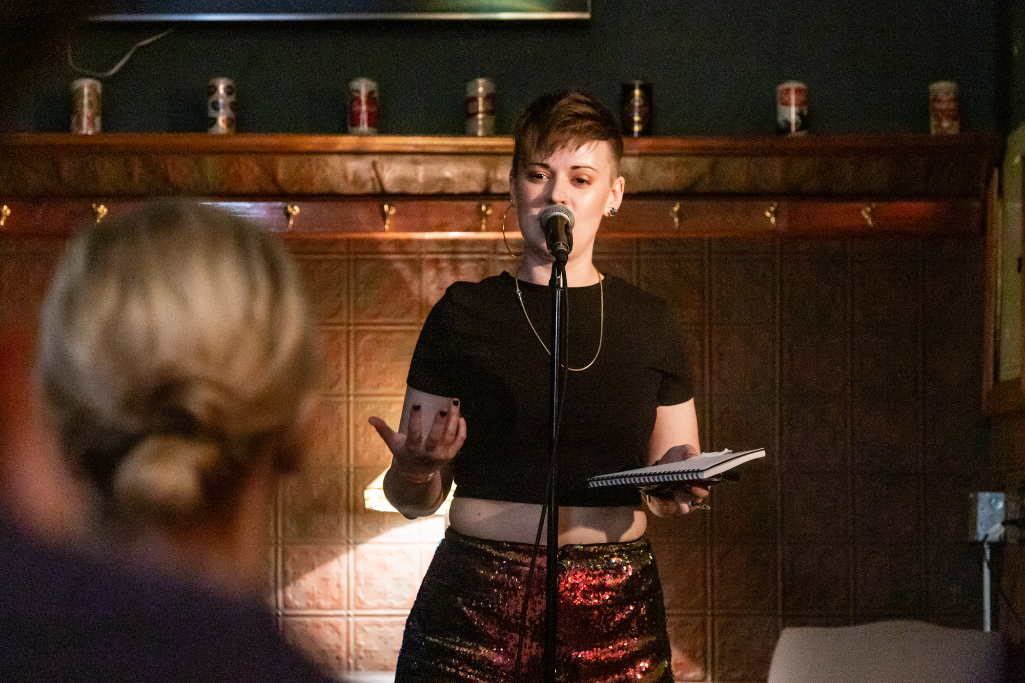 Featured image: Chelsea Fiddyment performing at Unreal at Schubas. Chelsea stands at the front of the room, notebook in hand, speaking into a microphone while looking out at the audience. Behind Chelsea is a copper-colored wall, made of a grid of low-relief tiles; above that are several decorative beer cans on a ledge and a dark green section of wall. Chelsea wears a black cropped tee and red and gold sequined shorts. The backs of some audience members’ heads are fuzzy in the foreground. Photo by Joshua Clay Johnson.