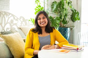 Featured image: Sharanya Sharma. Sharma sits with hands folded on a white table, with copies of “Set Fire to This Crooked House” and multi-colored notebooks in the foreground. Sharma wears a marigold cardigan open over a black and white striped shirt and smiles at the camera. Behind Sharma are several pastel throw pillows and a large plant, and natural light comes through the windows. Photo by Kristie Kahns Photography.
