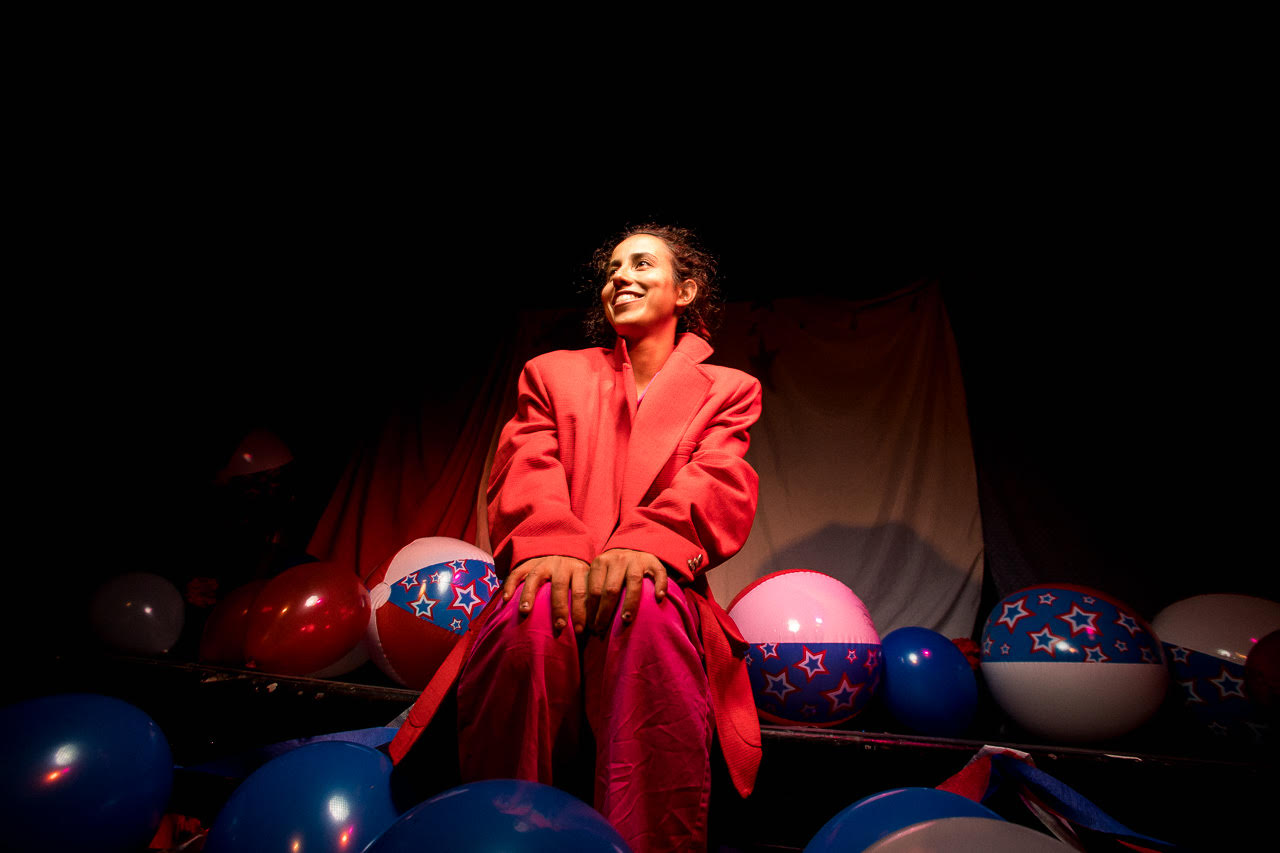 Image: Ida Cuttler, wearing a red blazer over pink pajamas, sits with her hands on her knees center. On the ground around her, are red and blue balloons and red-white-and-blue beachballs. The beachballs have stars on the blue stripes. Photo courtesy of Brave Lux, Inc.