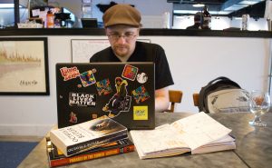 Image: Damian Duffy sits at a table in a brewery and works at a laptop computer. In front of his computer are paperback copies of Octavia Butler’s “Parable of the Talents”, an advanced reader copy of his graphic novel adaptation of Butler’s “Parable of the Sower”, and an open notebook with sketches. An empty beer glass is set to the side. Photo by Jessica Hammie.