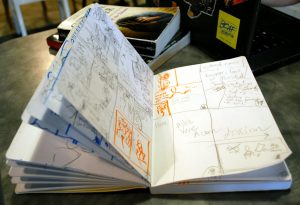 Image: A notebook sits on a table, open to pages filled with sketches and notes. Most sketches and notes are done in pencil, but there are a few that are done in orange marker. Photo by Jessica Hammie.