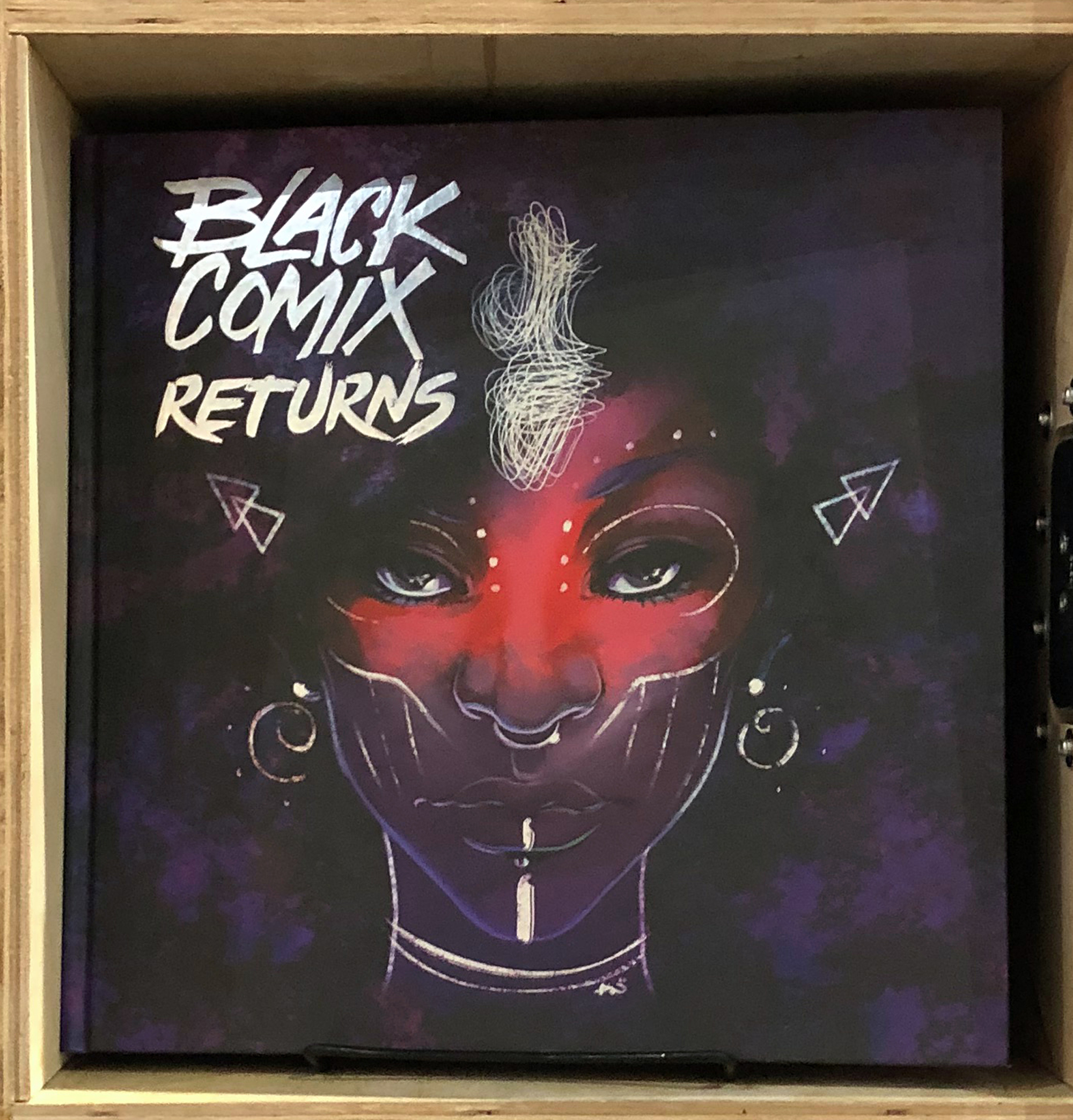 Image: Close up of “Black Comix Returns” on a wood bookshelf. The cover is various shades of purple and features a drawing of a woman’s face, with pink across the bridge of her nose and her cheeks. The artwork on the cover is by Ashley A. Woods. Photo by Jessica Hammie.