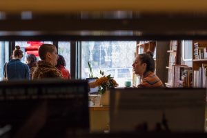Image: Framed from behind a bookshelf, a Pilsen Community Books customer (left) purchases a book from owner Mary Gibbons (right) at the front desk of the space. Photo by Amanda Dee.