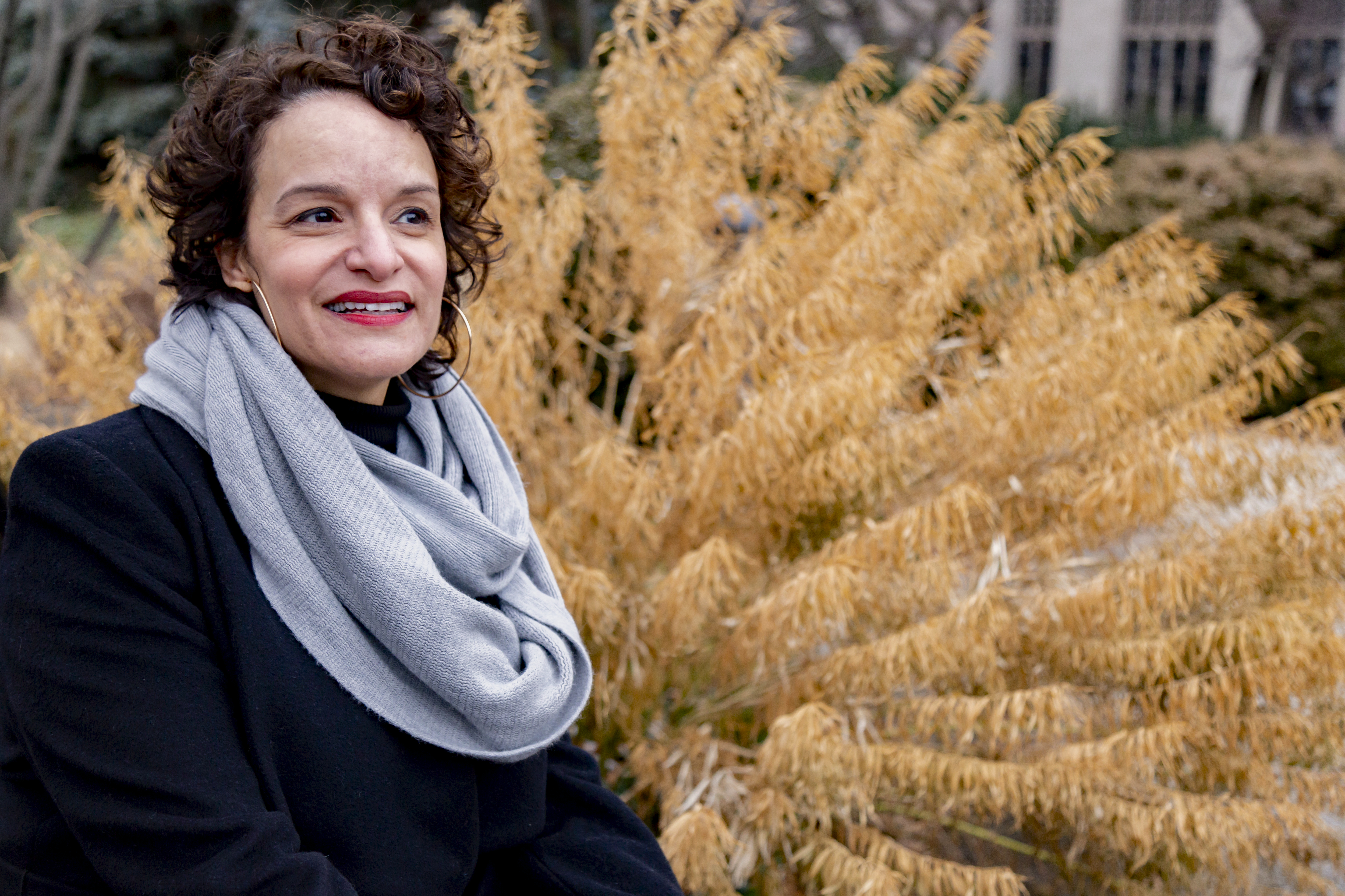 Featured image: Sahar Mustafah sits outdoors, smiling and looking off-camera. She wears a black winter coat, light grey scarf, and large hoop earrings. A bush with tan leaves fills most of the space behind her, with greenery and parts of a building behind that. Photo by Mark Blanchard.