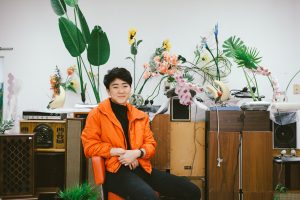 Rachel Youn in their studio. They sit in front of untitled works comprised of massagers, artificial plants, and speaker cabinets. Photo by Krista Valdez.