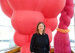 Featured Image: Amy L. Powell, Curator of Modern and Contemporary Art at Krannert Art Museum stands in front of “Hive,” an large inflatable sculpture installed in the museum’s Kinkaid Pavillion. The sculpture is floor to ceiling and bright pink. The main body of the sculpture resembles a bunch of grapes, or a multi-breasted female body, and to the side there is fuschia colored a braid with a braided gold band around the end of the braid. Powell stands in the center of the image, looking into the camera and smiling. She is wearing a black dress, and her hands are in her pockets. Photo by Jessica Hammie.