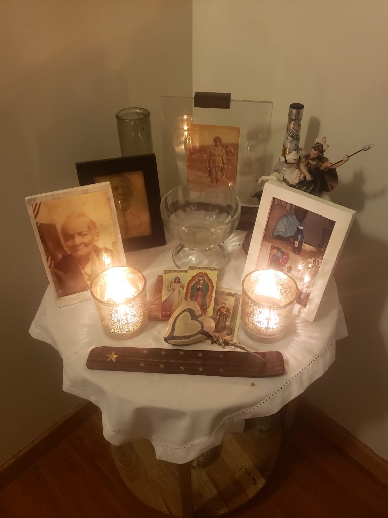 Image: Mario LaMothe's altar in his home. The altar consists of multiple framed photographs of LaMothe's family members sitting on a table with a white tablecloth. Candles and incense accompany the photographs. Photo courtesy of Mario LaMothe.