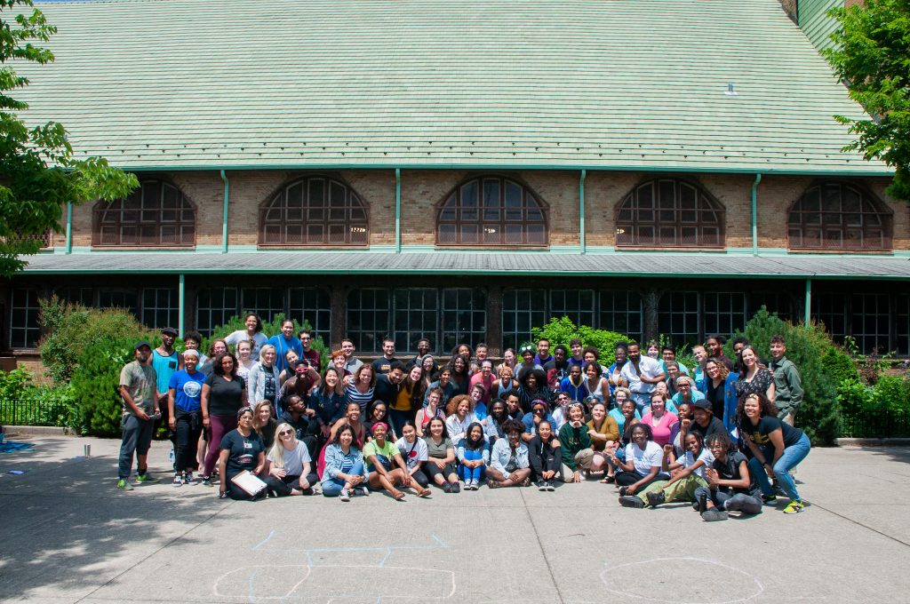  Image: The cast of Free Street Theater’s “50 in 50,” after dress rehearsal on June 22, 2019, outside at Pulaski Park. Several dozen performers crowd together — sitting or standing, many with arms around each other — and smile at the camera. The cast is multi-generational and predominantly BIPOC. Jagernauth kneels in the front row, farthest to the left; Spont-Lemus stands in the back row, near the center. Behind the cast is the backside of the Pulaski Park fieldhouse, and chalk drawings appear on the pavement in the foreground. Greenery peeks in from the edges of the frame and from behind the cast. Courtesy of Free Street Theater. Photo by Lena Jackson.