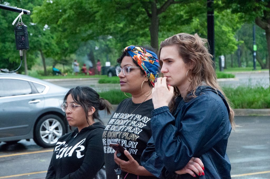 Image: Tanuja Devi Jagernauth (center) stands in a parking lot with Katrina Dion (right) and an audience member, intently watching an outdoor pop-up performance as part of “50 in 50” — Free Street Theater’s 50th anniversary celebration involving 50 performances in all of Chicago’s 50 wards in one day. They are positioned in near-profile, looking off-camera. Jagernauth wears a black t-shirt reading “WE DON’T PLAY,” glasses, and hoop earrings. The others wear dark-colored zip-up hoodies. A sound recorder hangs from a pole in the top left-hand corner of the frame, and the space farther behind the onlookers is lush and green. Courtesy of Free Street Theater. Photo by Lena Jackson.