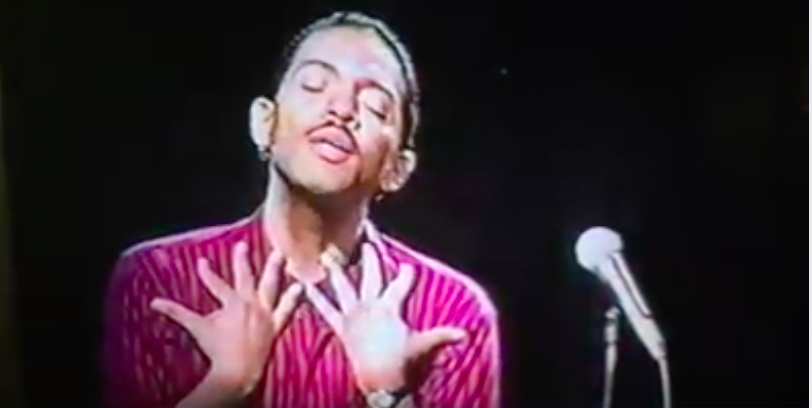 Image: A still from a video recording of Assotto Saint reciting his poetry. Assotto is in mid-word with his eyes closed while holding both hands to his body.