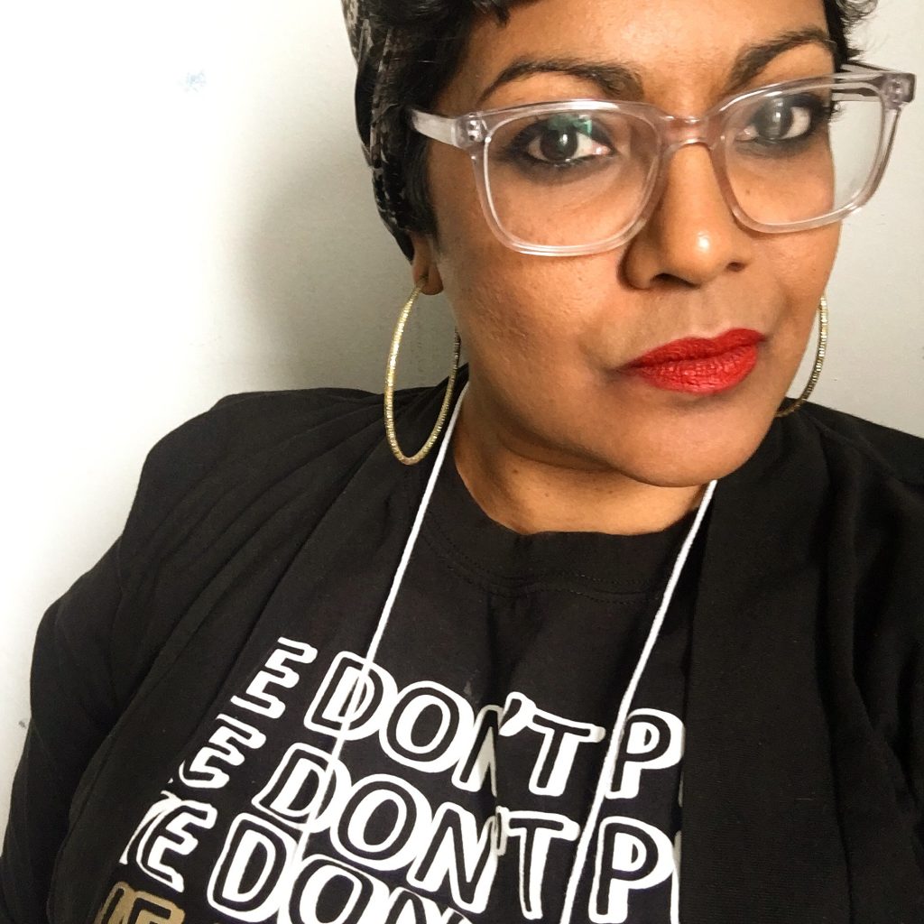 Image: Tanuja Devi Jagernauth. In this medium close-up photo, a self-portrait, Jagernauth looks at the camera with a firm expression. She wears glasses, red lipstick, hoop earrings, a headwrap, and a black cardigan over a black shirt that reads “WE DON’T PLAY,” repeated. Photo courtesy of the artist.