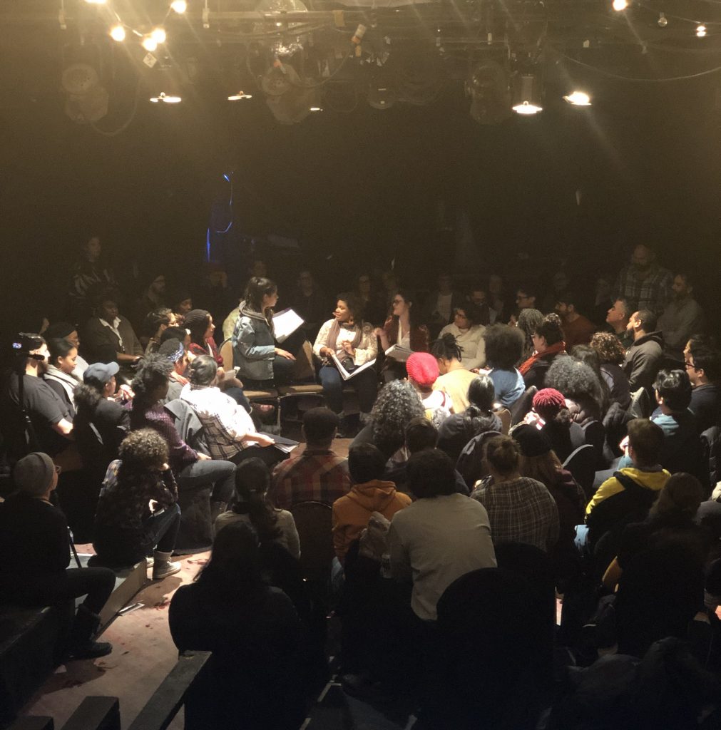 Image: A live reading of Tanuja Devi Jagernauth’s play, “How to Pick a Lock,” at Prop Thtr in 2019. In the center, a seated performer speaks to a performer who is crouched on a chair; other performers sit near them. All hold scripts. An audience of dozens encircles them. Stage lights and stringed lights hang from the ceiling. Photo by Tara Branham.