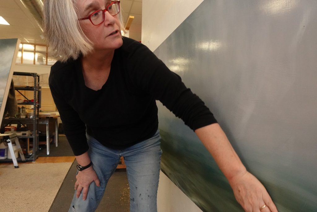 Image: The artist is leaning slightly forward wearing a black long-sleeved shirt and blue jeans. She is tilting her head towards the right of the photo with her hand working on a painting. Photo by David Travis.