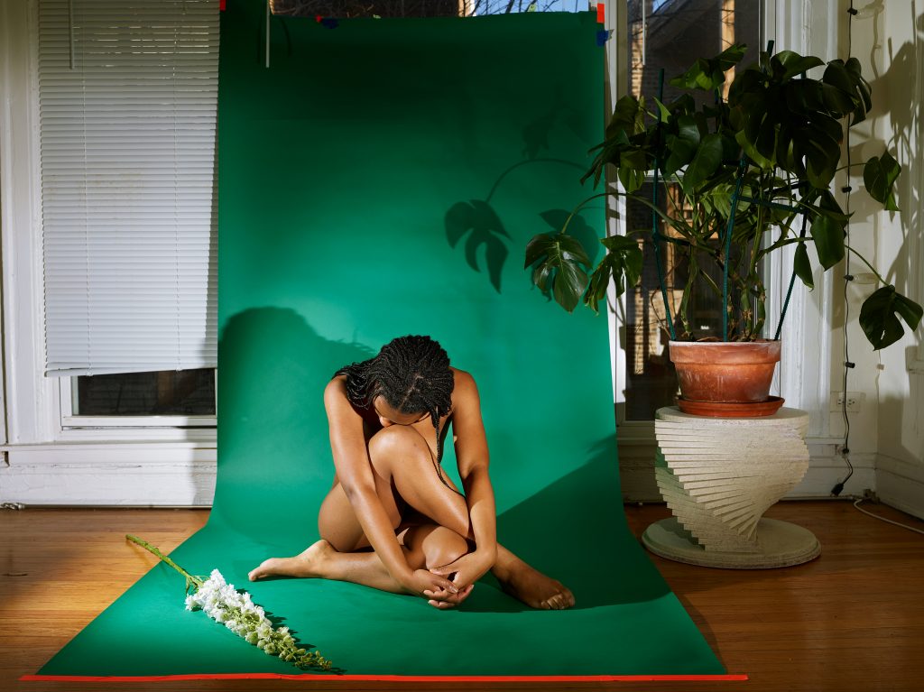 Image: Line by Marzena Abrahamik. A photograph of a Black woman sitting nude on the ground with a flower nearby. The person is sitting on a green sheet next to a larger plant. Image courtesy of the artist.