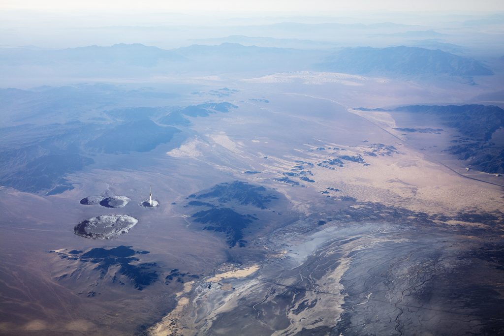 Image: Rocket Test, Mojave Desert, 1942/2019 by Barbara Diener. A color, aerial photograph of the Mojave Desert. There are crater-like shapes on the left-hand side of the photo. Image courtesy of the artist.