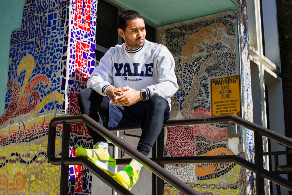 Image: Kristoffer McAfee is sitting on a metal railing in front of a doorway covered in colorful mosaic tile. He is wearing a Yale sweatshirt and look to the right with his hands together.  Photo by Kristie Kahns.
