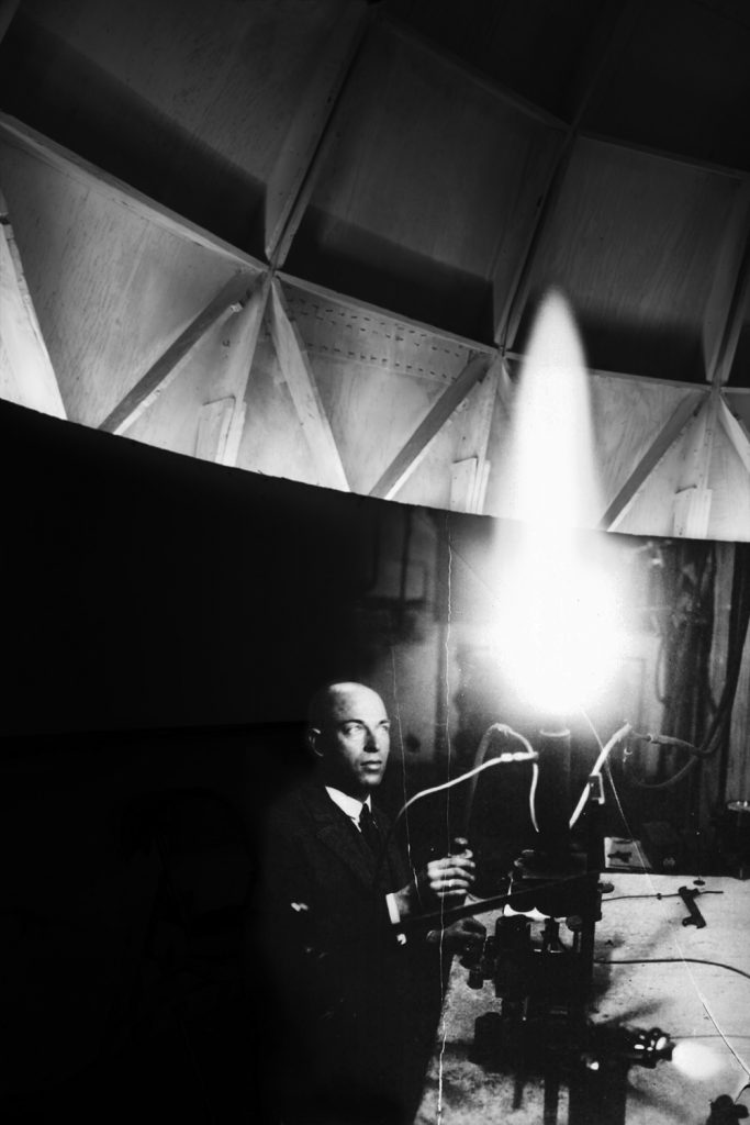 Image: Fuel Test, Peenemünde, 1939/2019 by Barbara Diener. A black and white photograph of a man conducting a fuel test. A bright light from the test is on the right side of the photo, with the left half dominated by darkness. Image courtesy of the artist.