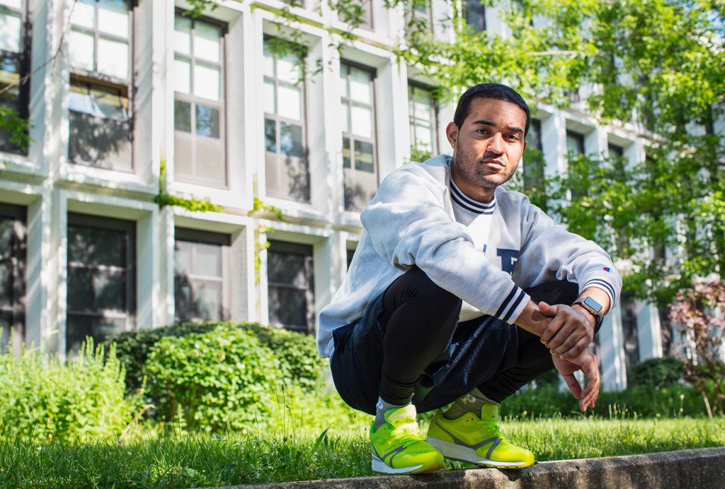 Image: Kristoffer McAfee in front of Kenwood Academy High School, a place where he says his interest in art was nurtured, and he emerged as a painter. McAfee is wearing a white sweatshirt, black pants, and neon yellow sneakers while squatting on a concrete ledge. He is looking directly at the viewer.  Photo by Kristie Kahns.