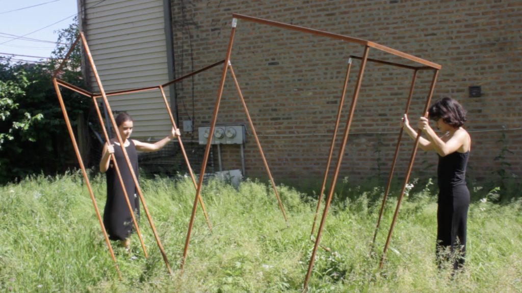 Image: "Join" by Cecilia and Marina Resende Santos, an installation/performance that took place on June 6, 2020 at an empty lot for sale by Landmark Property in Bridgeport. Two people pull apart an octagon metal frame. Photo by Graham Livingston.