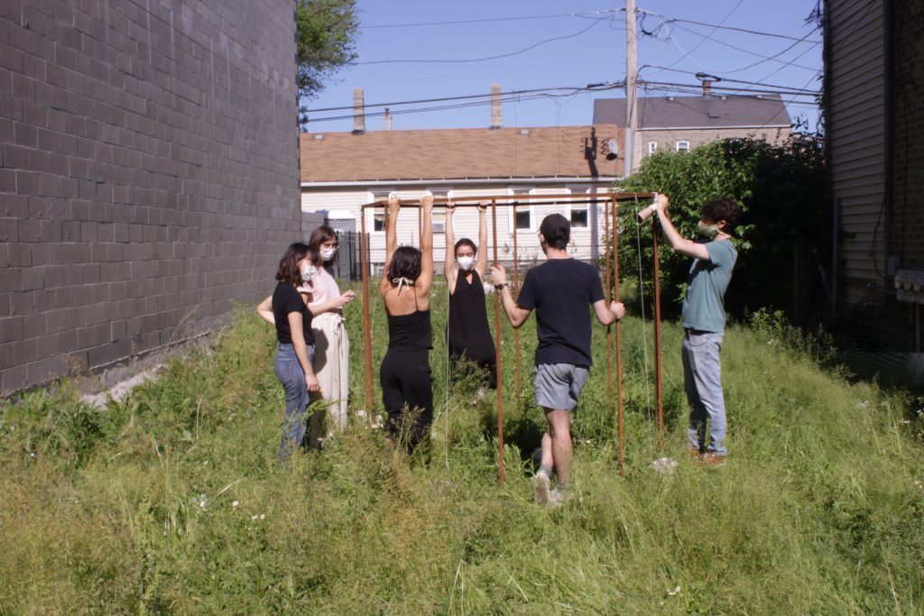 Image: "Join" by Cecilia and Marina Resende Santos, an installation/performance that took place on June 6, 2020 at an empty lot for sale by Landmark Property in Bridgeport. Six people stand around an octagon metal frame. Photo by Graham Livingston.