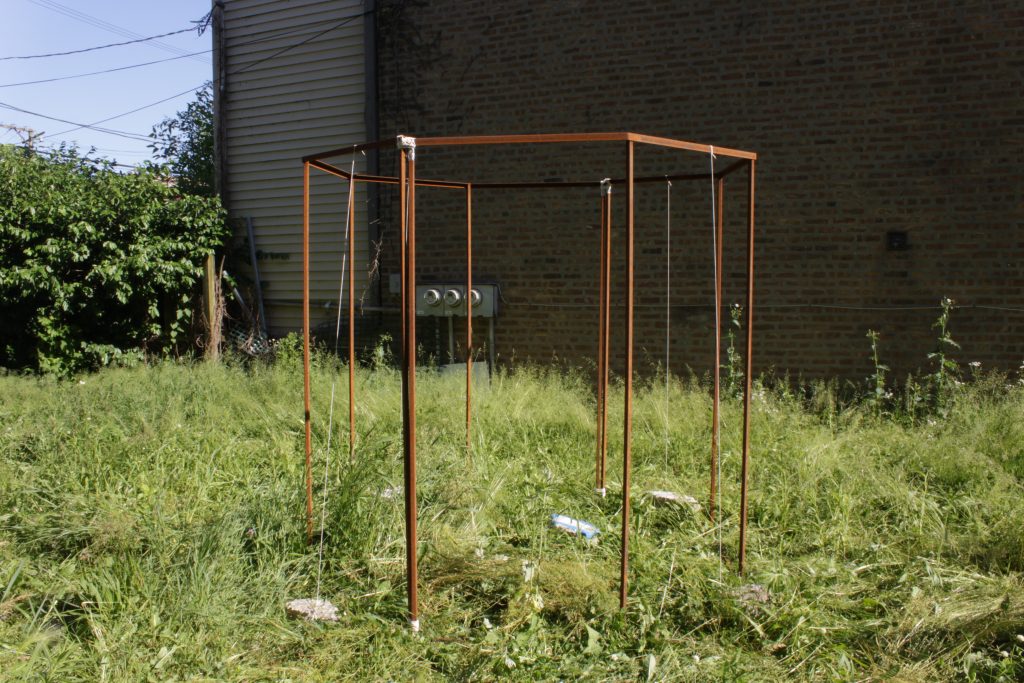 Image: "Join" by Cecilia and Marina Resende Santos, an installation/performance that took place on June 6, 2020 at an empty lot for sale by Landmark Property in Bridgeport. An octagon metal frame sits in a lot full of long grass. Photo by Marina Resende Santo.