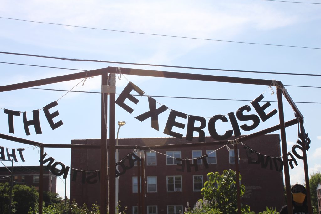 Image: "Untitled Politics" by Uniymeabasi Udoh, installed on July 13, 2020 at El Paseo Community Garden on Cullerton St and Csangamon St. in East Pilsen. Letter garlands are installed across the tops of an octagon metal frame. The letters in the foreground spell "EXERCISE". Photo by Cecilia Resende Santos.