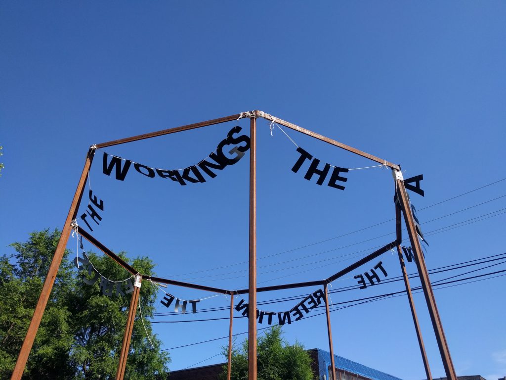 Image: "Untitled Politics" by Uniymeabasi Udoh, installed on July 13, 2020 at El Paseo Community Garden on Cullerton St and Csangamon St. in East Pilsen. Letter garlands are installed across the tops of an octagon metal frame. The letters in the foreground spell "WORKINGS" and "THE". Photo by Cecilia Resende Santos.