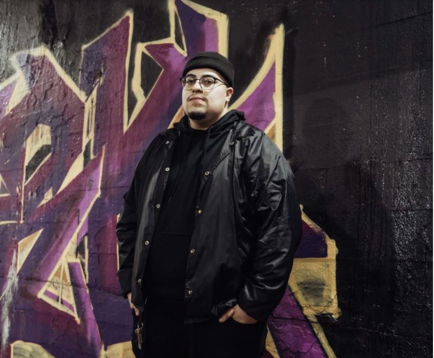 Featured Image: CeaseDays is wearing a black beanie, a black windbreaker with a black hoodie underneath, and black jeans. He has his hands tucked in his jean pockets. He is standing in front of a graffiti mural with a black background and purple and yellow letters.