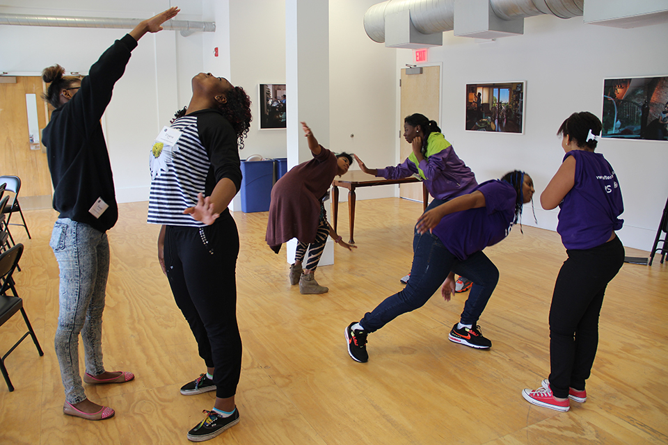 Image: Quenna Lené Barrett facilitating and participating in a Theatre of the Oppressed exercise with the Community Actors Program at the Arts Incubator in Washington Park, summer 2014. Barrett (center, wearing brown) and five teens, all young Black people, stand in pairs around a large room. One member of each pair uses their right hand to “lead” the other person’s face, such that one “follower” is standing tall and looking up to the ceiling and two “followers” are bent at the waist and leaning to the side. The performers wear casual clothing and two wear purple After School Matters t-shirts. Photos by Cecil McDonald Jr. are visible on the back walls. Photo by Marya Spont-Lemus. Image courtesy of University of Chicago Arts + Public Life.