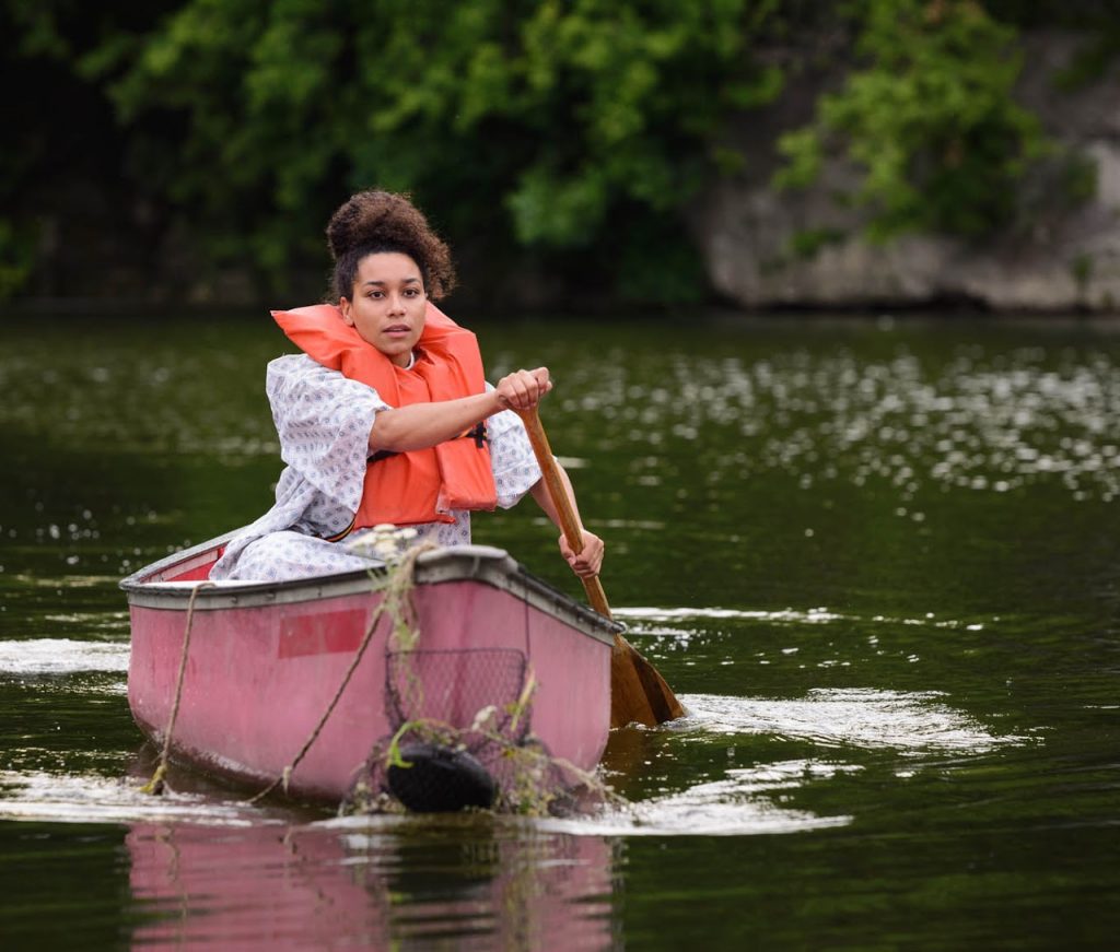 Image: A scene from “This Boat Called My Body.” In this wide shot of the outdoor performance, a young Afro-Latina woman (played by Elena Victoria Feliz) canoes toward the camera, through Palmisano Park’s quarry pond. She wears an orange life jacket and a white and blue patient gown. The pond water ripples from the performer’s paddling and the canoe’s movement. There is a stone quarry wall covered in greenery visible beyond that. Photo by Michael Courier. Courtesy of For Youth Inquiry and the Illinois Caucus for Adolescent Health.