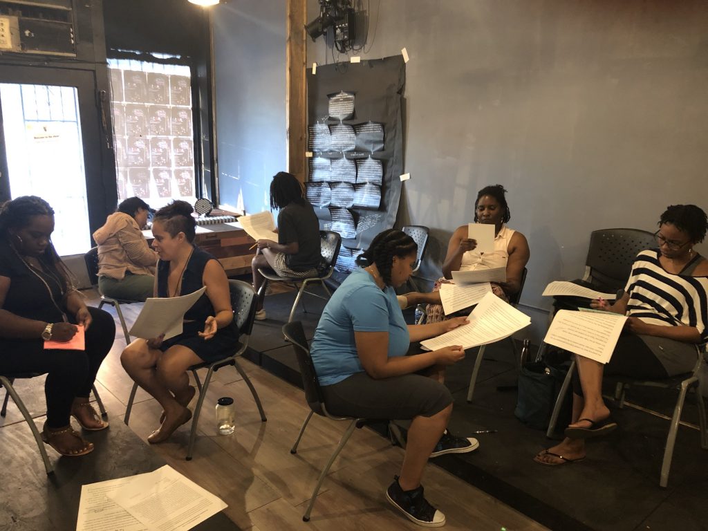 Image: A multi-day workshop for “Re-Writing the Declaration,” held at Free Street Theater’s Storyfront space in Back of the Yards, 2019. The photo shows a group of seven Black artists and artists of color seated in duos or trios around the storefront’s interior. From left to right: Teharah J. Smith, Kristiana Rae Colón, Deanalís Resto, Kirsten Baity, Zanneta Kubajak, Sharaina L. “Shay” Turnage, and Kanomé Jones. Each person appears to be discussing, taking notes on, and/or reading a copy of the same document, printed in black ink on large white paper. One wall is grey and the other is windowed, with light coming in from outside. A piece of black butcher paper is taped to the far wall, with white-on-black printings attached to it. Photo by Quenna Lené Barrett.