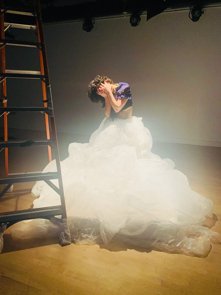 Image: A behind the scenes look at the performance Four Walls and One Me by Taimy Ramos Velázquez. A dancer stands with hands over face wearing a large white skirt.  A ladder stand to the left. Photo courtesy of the artist.