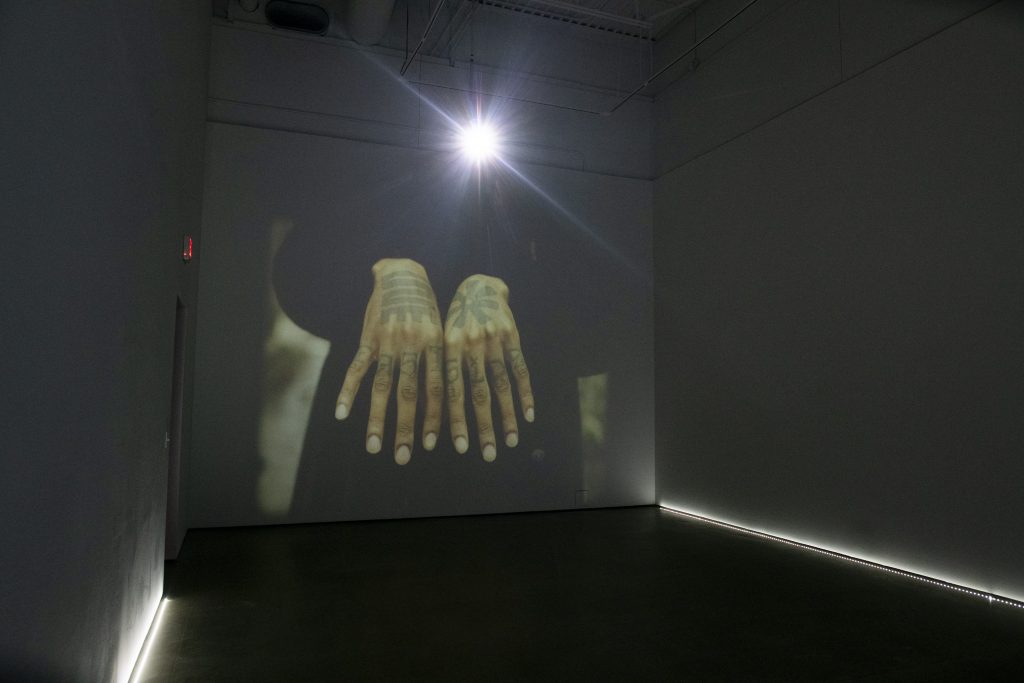 Image: Installation view of The Line by Cameron Granger. The video piece is being projected on a large wall. The still being shown is of a person holding out their hands palms down, showing tattoos on their hands. Image courtesy of the gallery. Photo by Ty Wright.