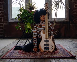 Image: A photo of Tonina Saputo sitting on a rug while posing with her guitar. Photo by Danny Zones, courtesy of the musician and the photographer.