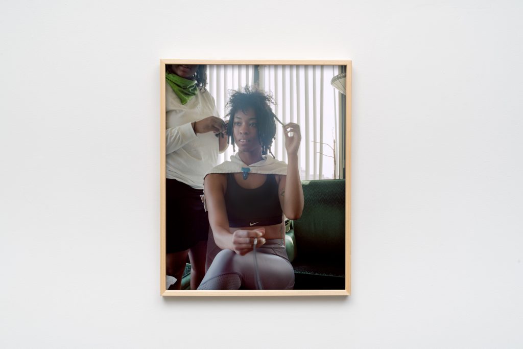 Image: Jazmine Harris, Untitled, 2020, archival pigment print. The artist is center frame, her gaze above and to the viewer’s left. The artist sits at home in her grandparent’s chair while she has her hair done. In her hand is a remote shutter release cable. Photo courtesy of the artist and curator.