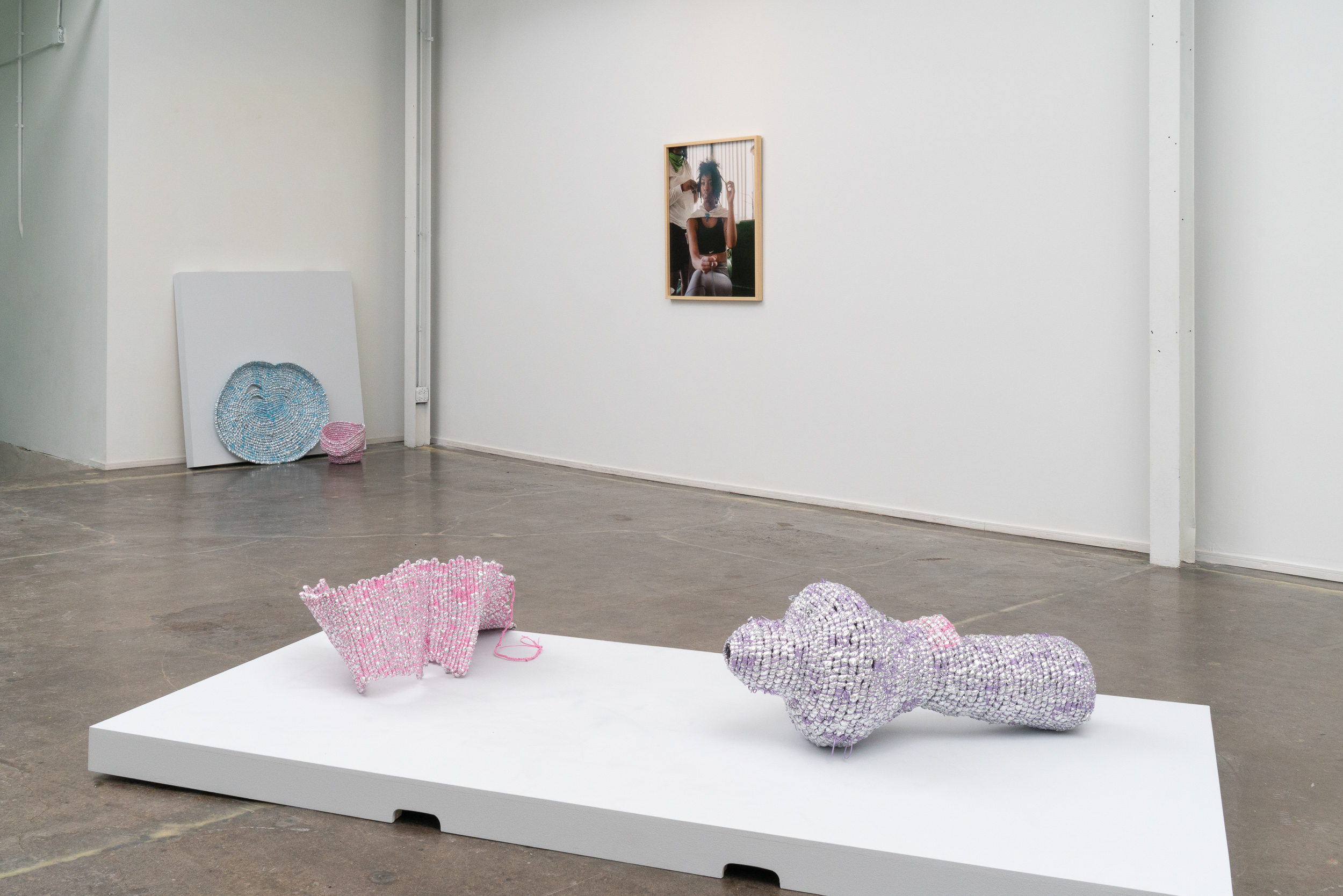 Image: Installation view of Chicago Artists Coalition’s Repository and Repertoire. José Santiago Pérez's sculptural pieces Un/Burden (so you may release), 2020, and Un/Burden (so you may restore), 2020, sit in the foreground on a white exhibition plinth. The piece on the left is composed of light-pink coiled emergency blankets and plastic lacing. The piece on the right is composed of silver and purple emergency blankets and plastic lacing. In the background Jazmine Harris' self-portrait Untitled is exhibited on the wall. To the left of Harris' piece, is Perez's, Un/Burden (so you may continue), 2020: two rimmed sculptural light pink and aqua-marine sculptural forms composed of the same materials. Photo courtesy of the artist and curator.