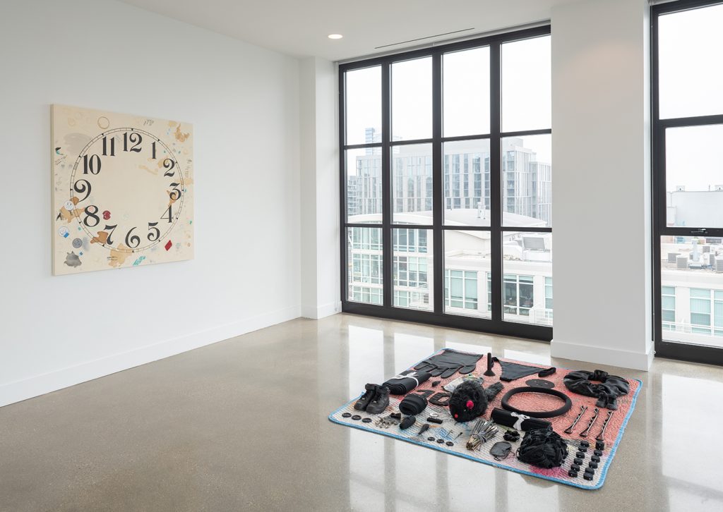 Image: Amanda Ross-Ho, installation view of Usual Objects. (l) Untitled Timepiece (ORAL ARGUMENTS/OVERLAPPING JURISTICTIONS), 2019-2021. Gesso, silkscreen, acrylic, ink, ball point pen, graphite, oil pastel, coffee, tea, paper, artist’s tape, painter’s tape, laser prints, stretch sequin fabric on canvas covered panel, 52 x 52 x 1.5 inches. (r) Untitled Seizure (ENCORE/DARK MATTER), 2021. Unfinished Black Glove (small), empty thread spool (UNTIRLED PERIOD PIECE), Extra Thong (ONCE YOU GO BLACK), facemasks, Giant Scrunchie (MY PEN IS HUGE), Cut and bundled pant legs (Untitled Production Line (Modern Times), black mouse, BLACK GLOVE FINGERTIP, Mouse (Yin), BINARY (Rubber Band), BINARY (Bobby Pin (3)), 10, Untitled Metrics (TENS), cut and bundled pants parts (Untitled Production Line (Modern Times),Nike Air Force Ones (UNTITLED PERIOD PIECE), Bundled and pinned canvas (Untitled Production Line (Modern Times), Black Earring (Untitled Peripheral Swatch (FOOTPRINTS AND FREE RADICALS)), pinned canvas (Untitled Production Line (Modern Times), Dead Brushes (Periphery Composition with Basket of Dead Brushes), satin scrunchie, Delta eyemask, canvas spaghetti (Untitled Production Line (Modern Times), vintage clock numbers, pinned canvas pucks (Untitled Production Line (Modern Times), Buttons Large (Untitled Production Line (Modern Times), Black Nitrile Glove, Rolled canvas (Untitled Production Line (Modern Times), Black 3D Printed Plastic silverfish (Untitled Production Line (Modern Times), Clock hands (Untitled Production Line (Modern Times), Buttons Small (Untitled Production Line (Modern Times), 77 x 67 x 5 inches. Image courtesy of the artist and Carrie Secrist Gallery.