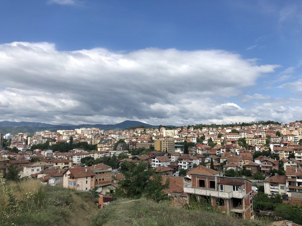 Image: A photo overlooking half of the city of Sandanski, Bulgaria. The edge of the cliff on which the picture is taken is visible in the bottom left. Buildings take up most of the lower half of the photograph - all with the traditional brick roofs. In the horizon are visible mountain ranges on the left and a row of trees on the right. Photo by the author.