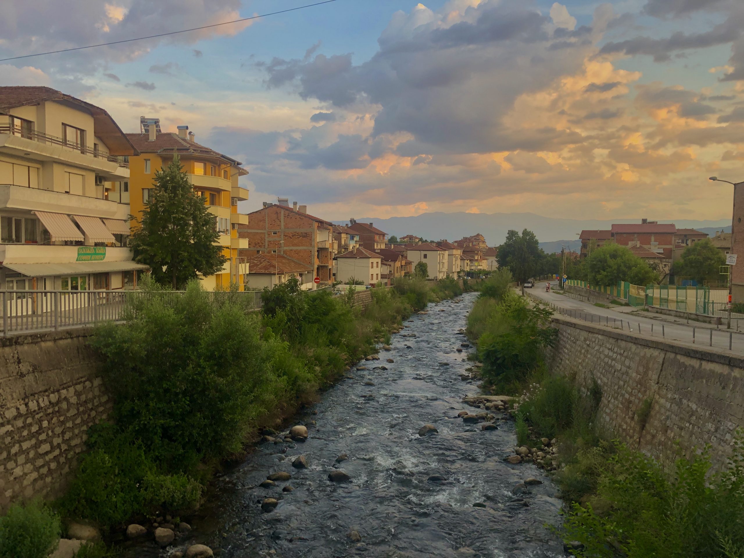 Featured image: A photograph of the Struma river, which cuts through the middle of the image (and also through the middle of Sandanski, Bulgaria), flanked by levees on either side. The water level is pretty low, so there is vegetation growing along the levees. On the left side of the river is a street and apartment complexes. On the right there is a street beyond which the edges of a school yard are visible. There is a mountain range visible in the distance. Photo by the author.