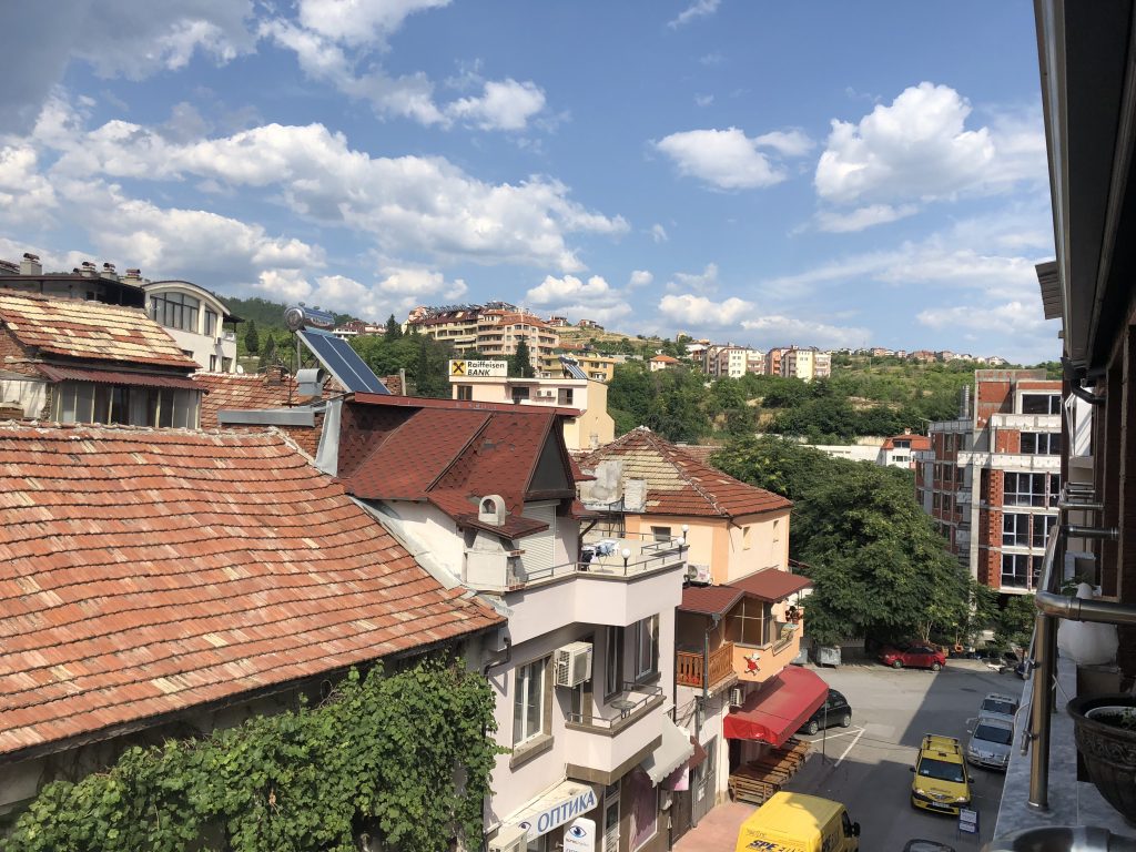 Image: A photo overlooking a small section of Sandanski as seen from the terrace of an apartment. The traditional brick rooftops are on the left.. The facade of the first building from the left is obstructed entirely by grape vines. The second building is white and has a sign that says "ОПТИКА," roughly translated to "optics" (it is an optometrist office). The third building is a peach color and the first level appears to be a small outdoor cafe. There are cars parked on the street below. There is a building still under construction on the right - only the brick framework is complete. Lots of greenery and trees break up the individual buildings and it's a bright summer day. Photo by the author.