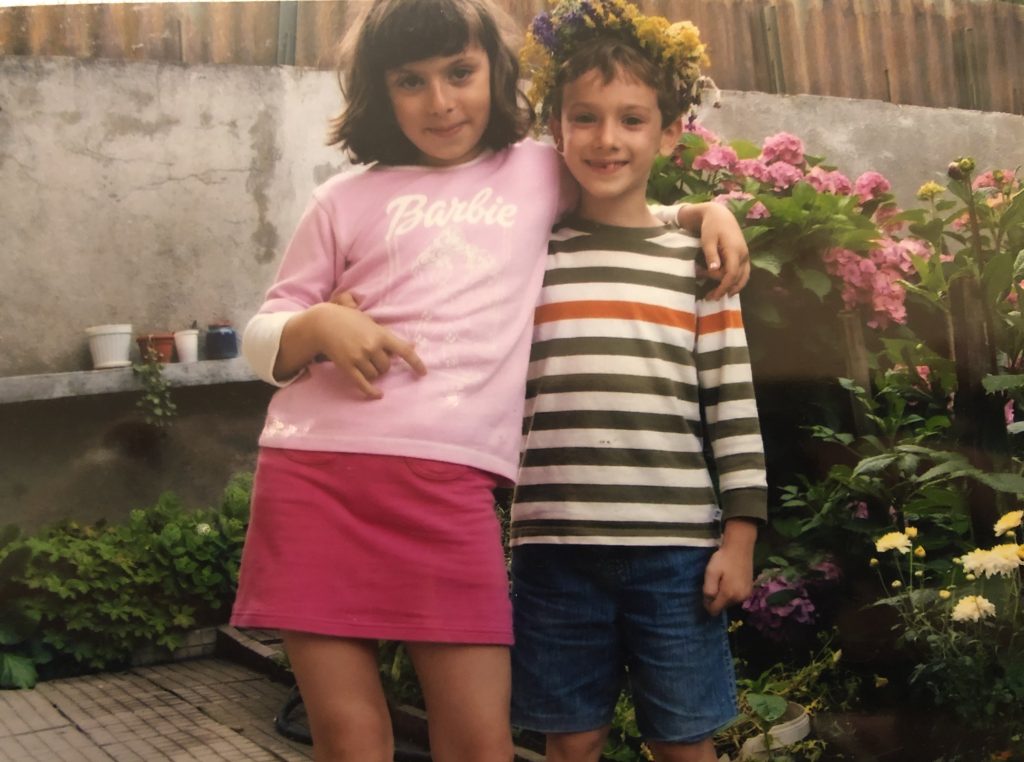 Image: A photo in a photo album of the author and her brother when they were children. They are standing in a sideways embrace in the yard of their grandmother's house. There is a stone wall behind them and lots of flowers on the right. The author wears a light pink "Barbie" long sleeve shirt and a magenta skirt. She has one hand slung around her brother. She has shoulder length brown hair and smiles at the camera. Her brother also smiles and is wearing a flower crown comprised of yellows and purples.. He wears a horizontally striped green, white, and orange long sleeve shirt and jean shorts. One hand is around his sister. Image courtesy of the author.