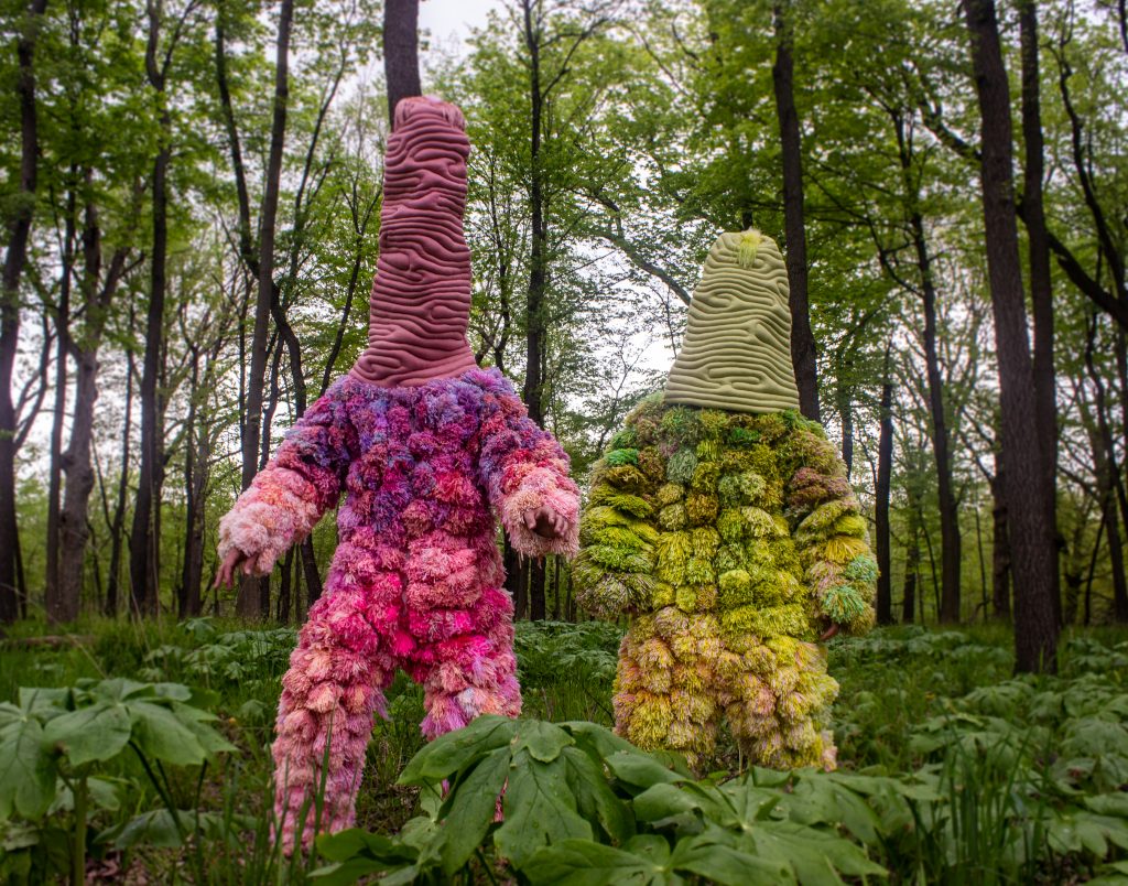 Brendan O’Shaughnessy, Microorganisms, upcycled and new yarn, upcycled fabric, feathers, fiberfil, and mixed media, 2020. Two figures wear upholstered, colorful, full-body suits with knit fabric hoods. They are standing in a lush wooded area. The figure on the left is pink and purple; the figure on the right is mostly yellow and green. Photo courtesy of Brendan O’Shaughnessy. 