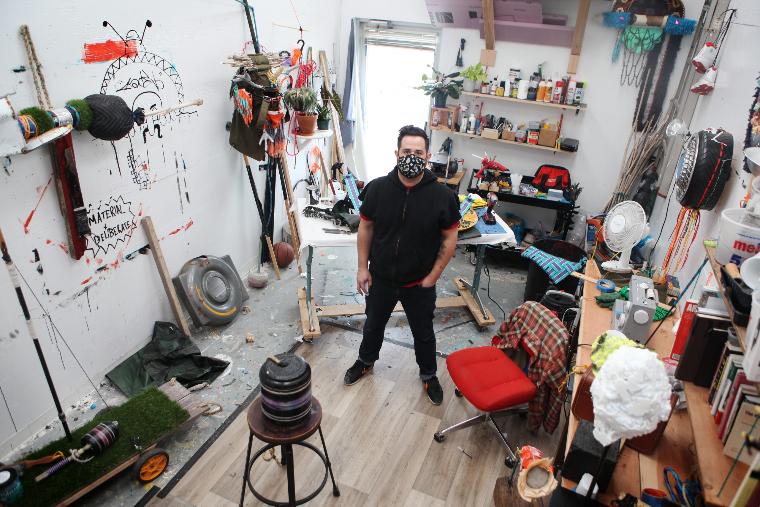 Featured Image: Stephen Signa-Aviles stands in his studio at the University of Illinois at Urbana-Champaign. He is wearing dark jeans and a dark, short-sleeved hooded sweatshirt, and a black face mask with a white graphic pattern. He looks up and into the camera. His studio space is narrow and cluttered. There are various works in progress, as well as shelving units with paints, books, and other materials. Photo courtesy of Stephen Signa-Aviles.