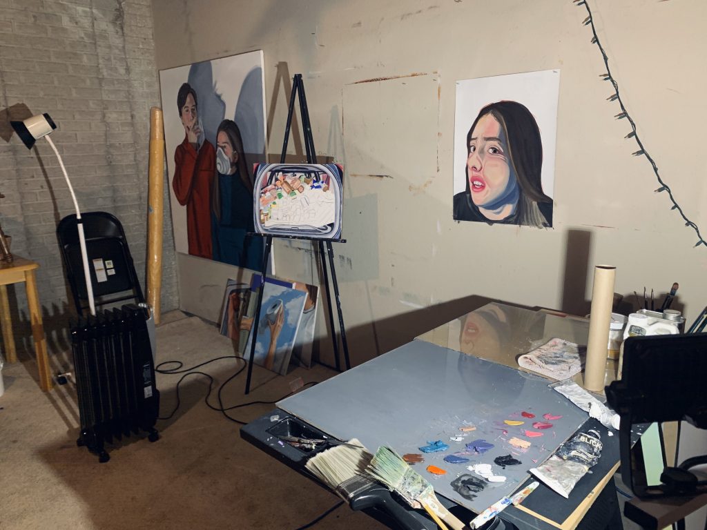 Image: A view of Pascale Grant’s studio work space in the basement of her home. On a neutral, drywalled basement wall are two paintings. There is a small stack of paintings on the floor leaning against the wall. A painting in progress is on an easel between the two wall-hung works. On the right of the image is a table with paint palettes, brushes, and other painting supplies. On the left of the image are a space heater, a tall lamp, and a folding chair leaning on an unfinished wall. Photo courtesy of Pascale Grant.