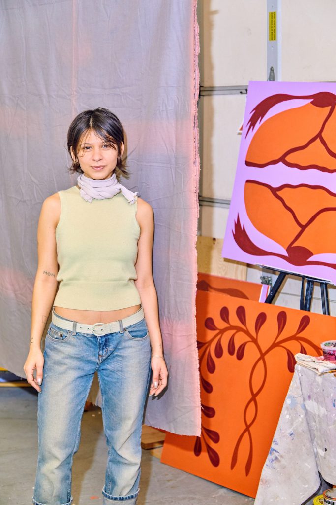 Image: Marissa Macias faces the camera while standing in front of a pale purple and pink cloth. She is wearing jeans, a white belt, and a pale yellow shirt. Several paintings sit in the background in her studio. Photo by Sarah Joyce.