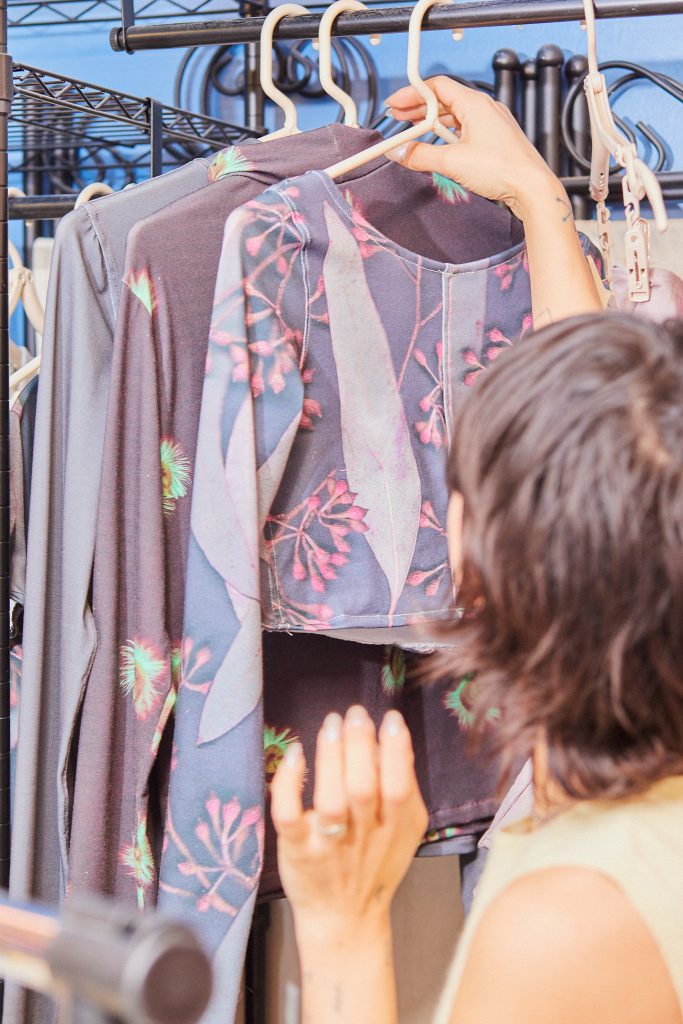 Image: Marissa Macias faces away from the camera looking at her clothing. Marissa is hanging a top on a rack where several other tops hang. The tops are mostly pale purple. Photo by Sarah Joyce.